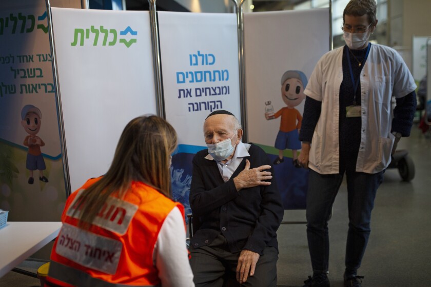 An elderly man wearing a skullcap speaks with a woman seated across from him as another woman stands next to him.
