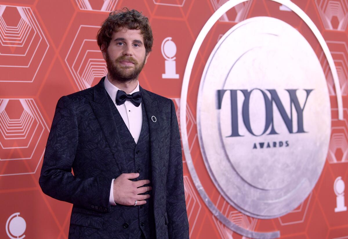 A man with scruffy brown hair and facial hair in a tuxedo against a red backdrop