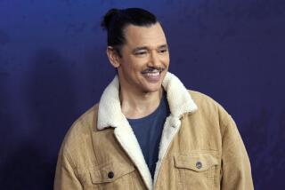 A man with black hair pulled up in a bun and a mustache smiling in a beige jacket and blue shirt