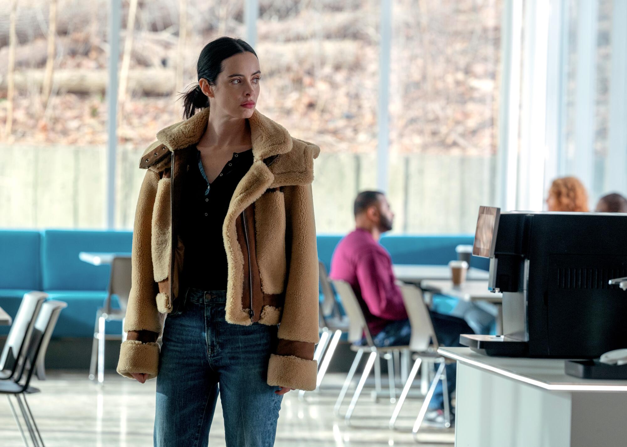 A woman in a brown aviator coat and jeans stands in a cafeteria.