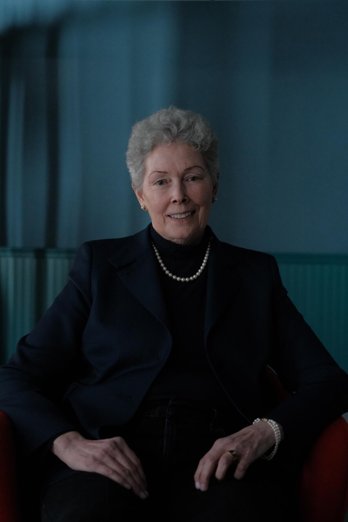 A woman with short gray hair on a dark turtle neck and a sitting blazer.