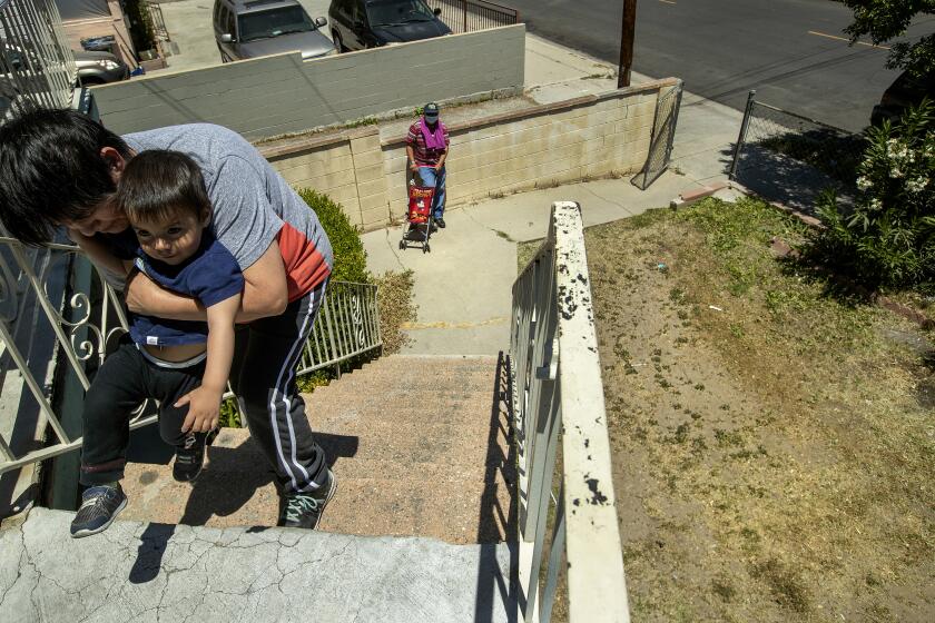 LINCOLN HEIGHTS, CA-MAY 11, 2022: Benita Perez, 55, teaches her grandson Nevaeh Perez, 1 and 1/2, how to walk at their apartment building on Thomas St. in Lincoln Heights. Area at right, below used to be all grass until a year ago, due to the drought. It's going to be a summer of brown grass and hard choices for Southern California lawn owners facing the Metropolitan Water District's one day a week watering restrictions starting June. 1. (Mel Melcon / Los Angeles Times)