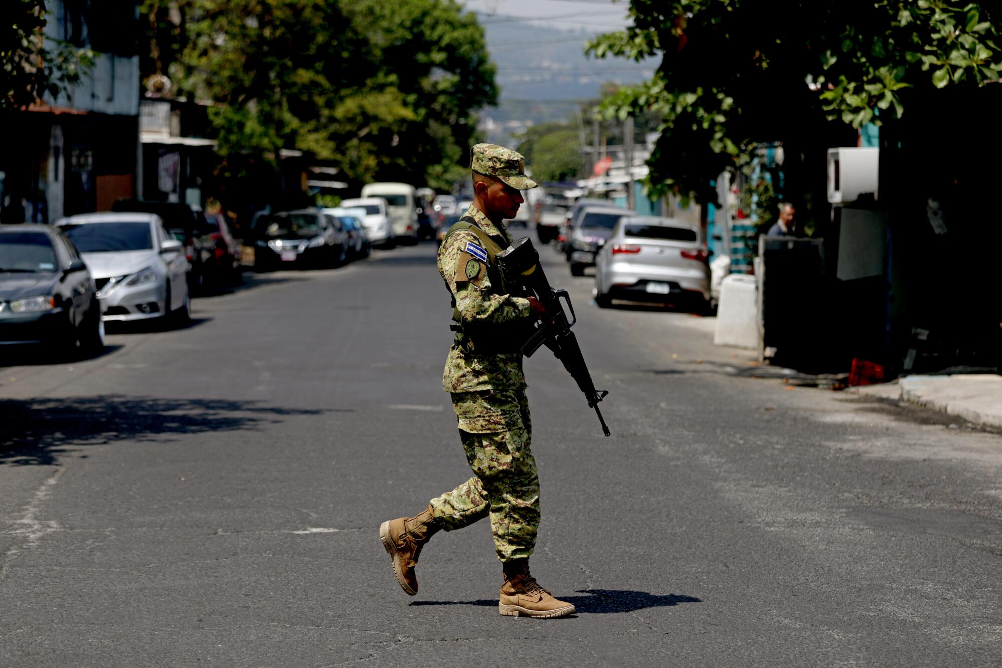  A person in fatigues, holding a gun,  walks across a street lined with cars