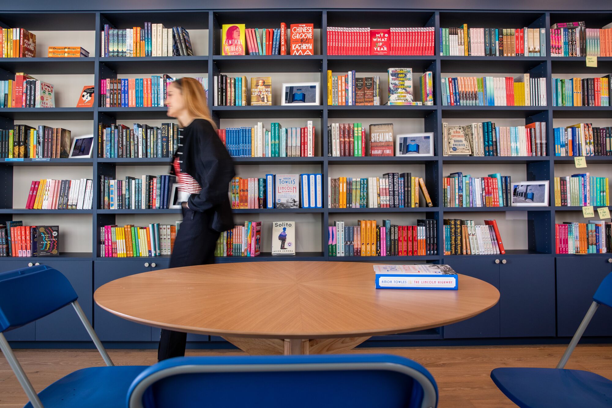 A woman walks between shelves of books and a round table with folding chairs around it.