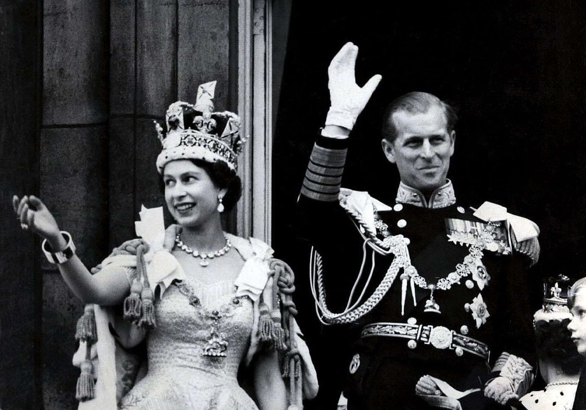 A woman wearing a crown and tasseled cloak, left, and a man in a military uniform with medals smile as they wave 