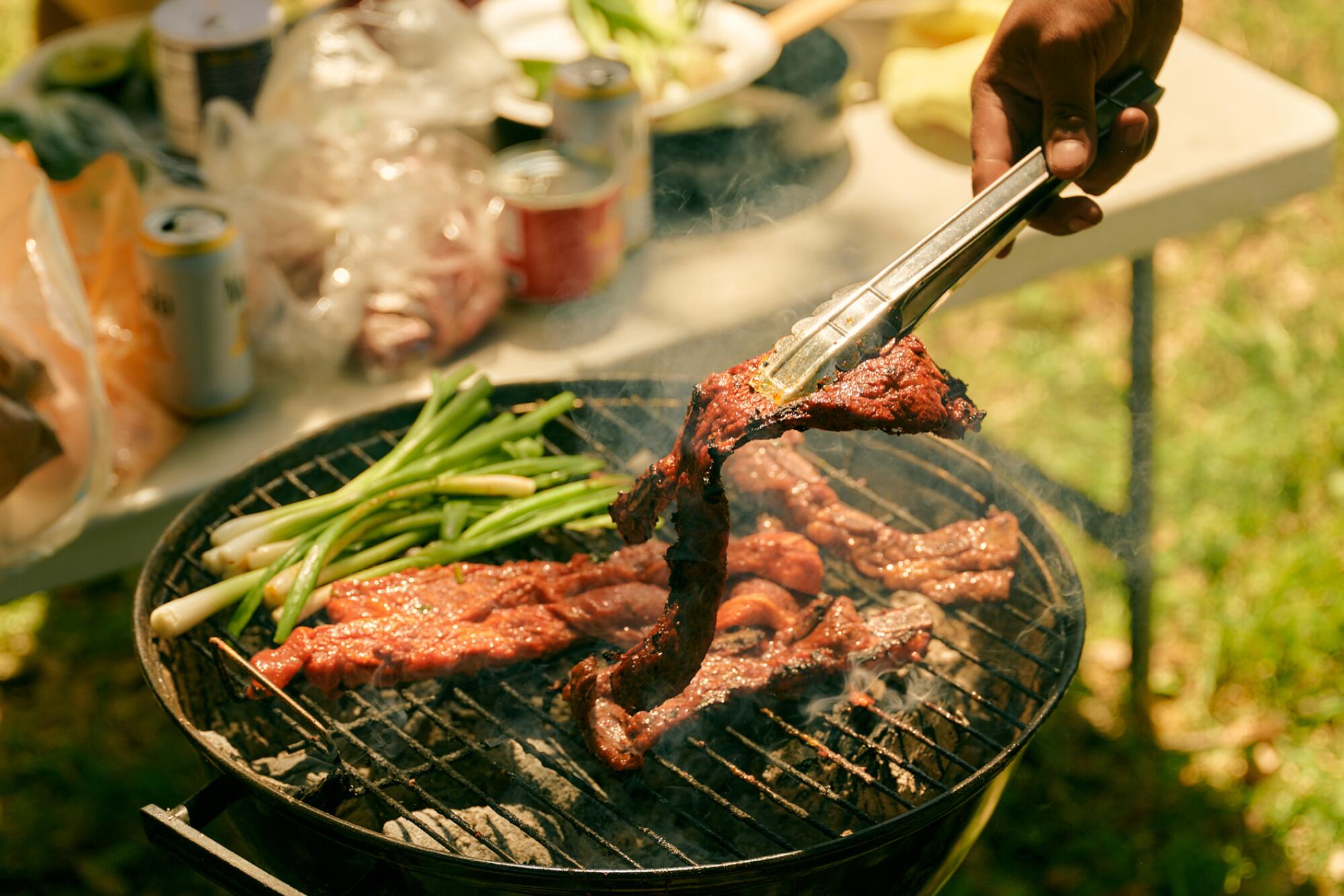 Tongs pick up one of the pieces of carne asada on a grill alongside scallions