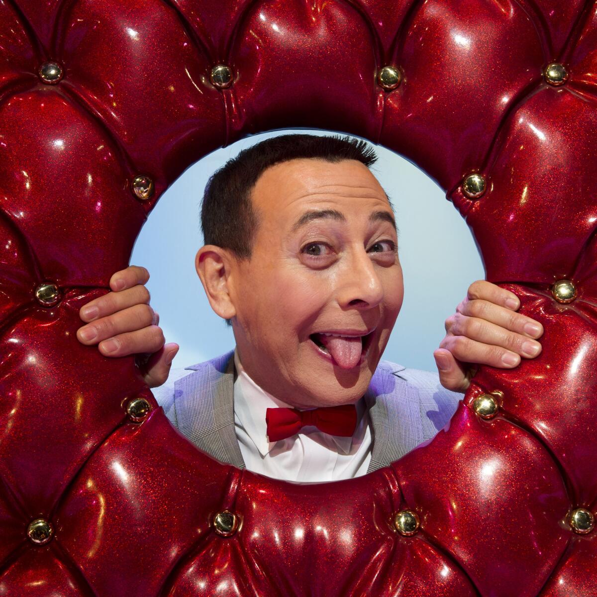 Paul Reubens sticks out his tongue while poking his head and hands through a hole in an upholstered red donut