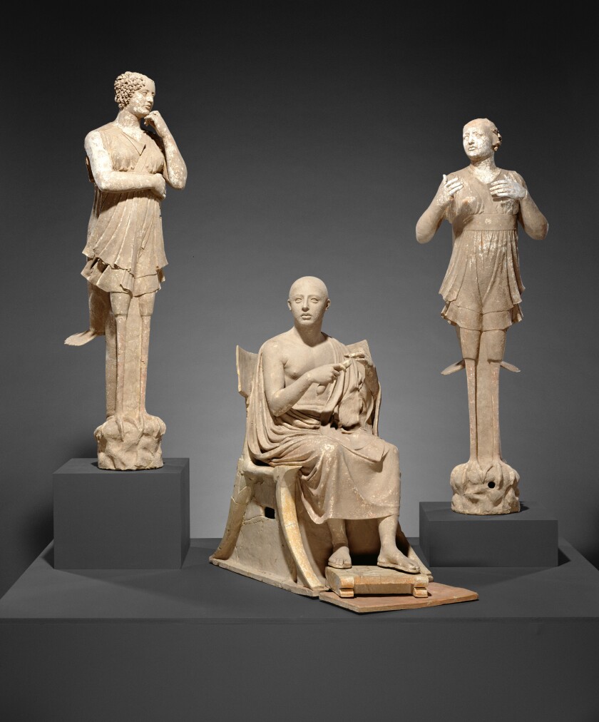 Ancient Greek statues of two women standing and a man sitting on a chair.