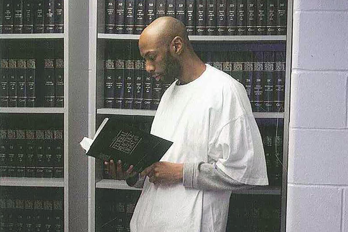 Dustin Higgs holds a book while standing in front of shelves filled with books.