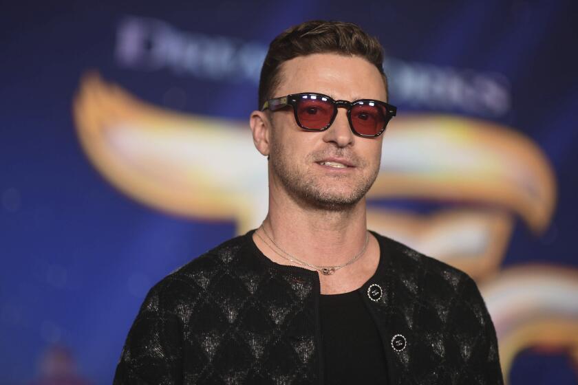 Justin Timberlake smiling. He wears sunglasses, a dark jacket and a black t-shirt and poses against a blue background