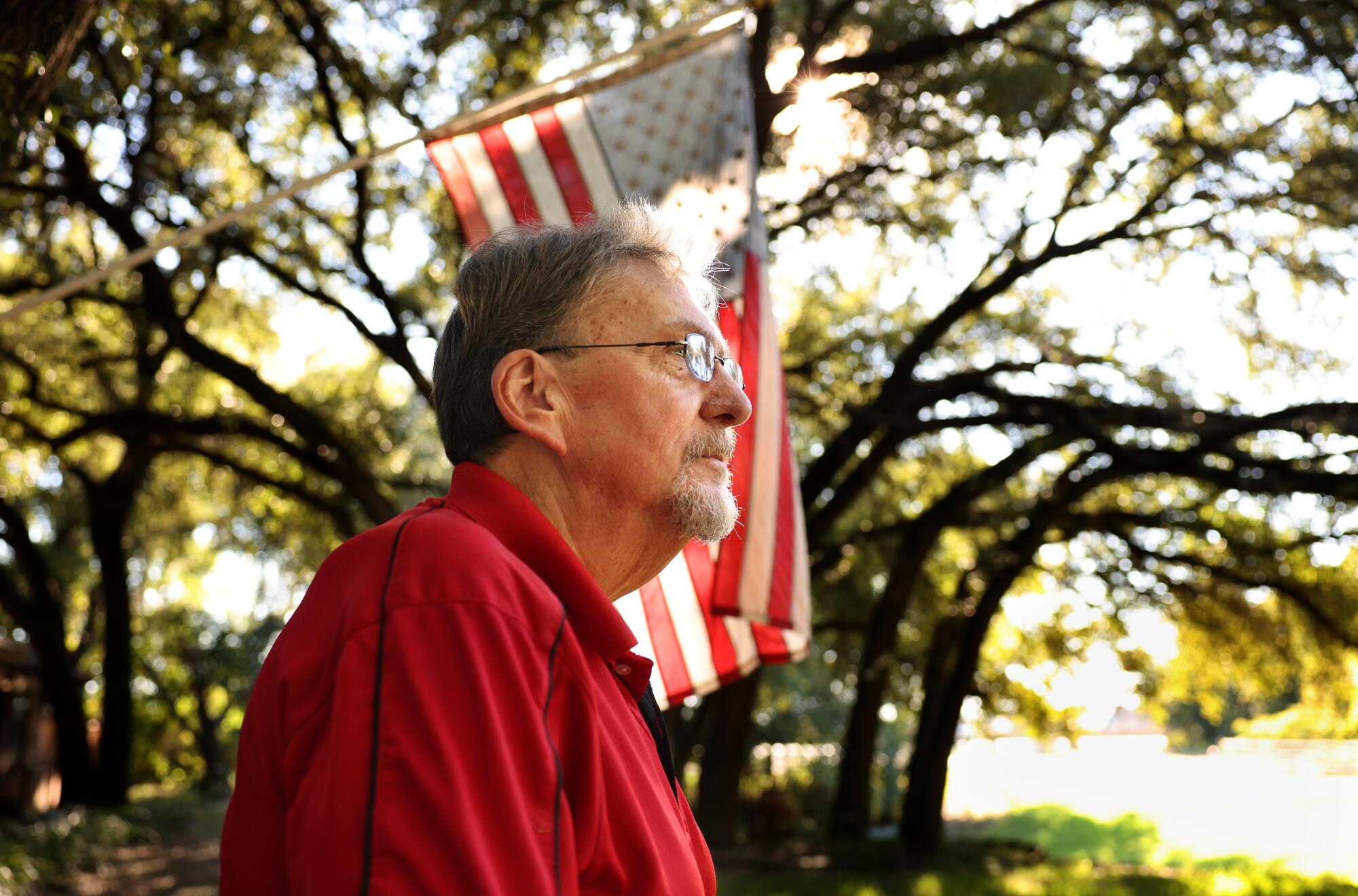 A man in profile wearing a red jacket and standing in front of an American flag with large trees in the background 