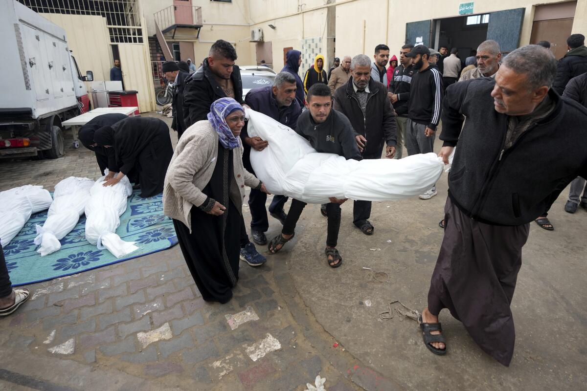 People carry a body wrapped in a white shroud, as a woman in black leans over three white-shrouded bodies on the ground