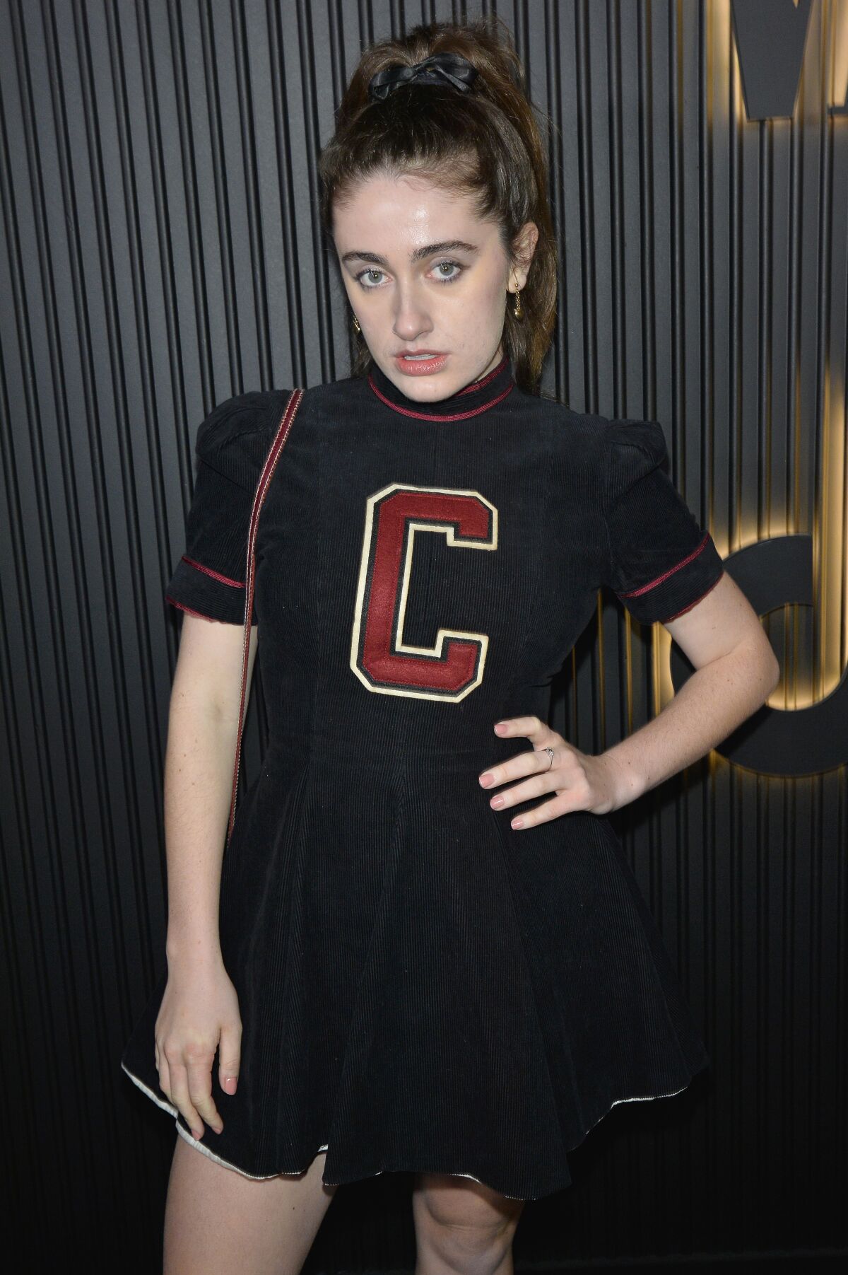 Rachel Sennott in a black outfit with a red C on the top and one hand on her waist.
