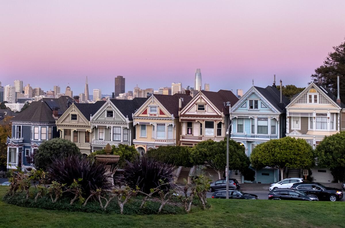 A row of pastel-hued Victorian houses line a street. San Francisco skyline is visible behind them.