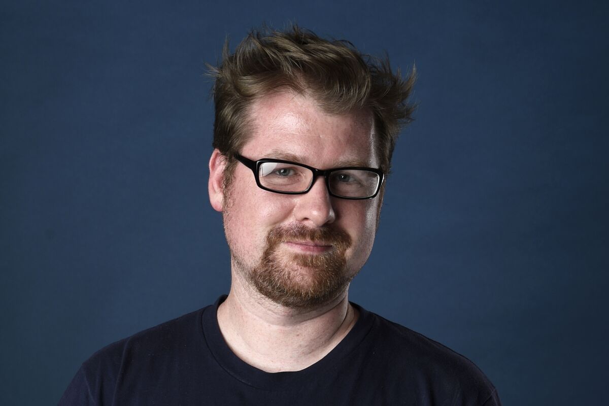 A man with facial hair and glasses smiles in front of a dark blue background