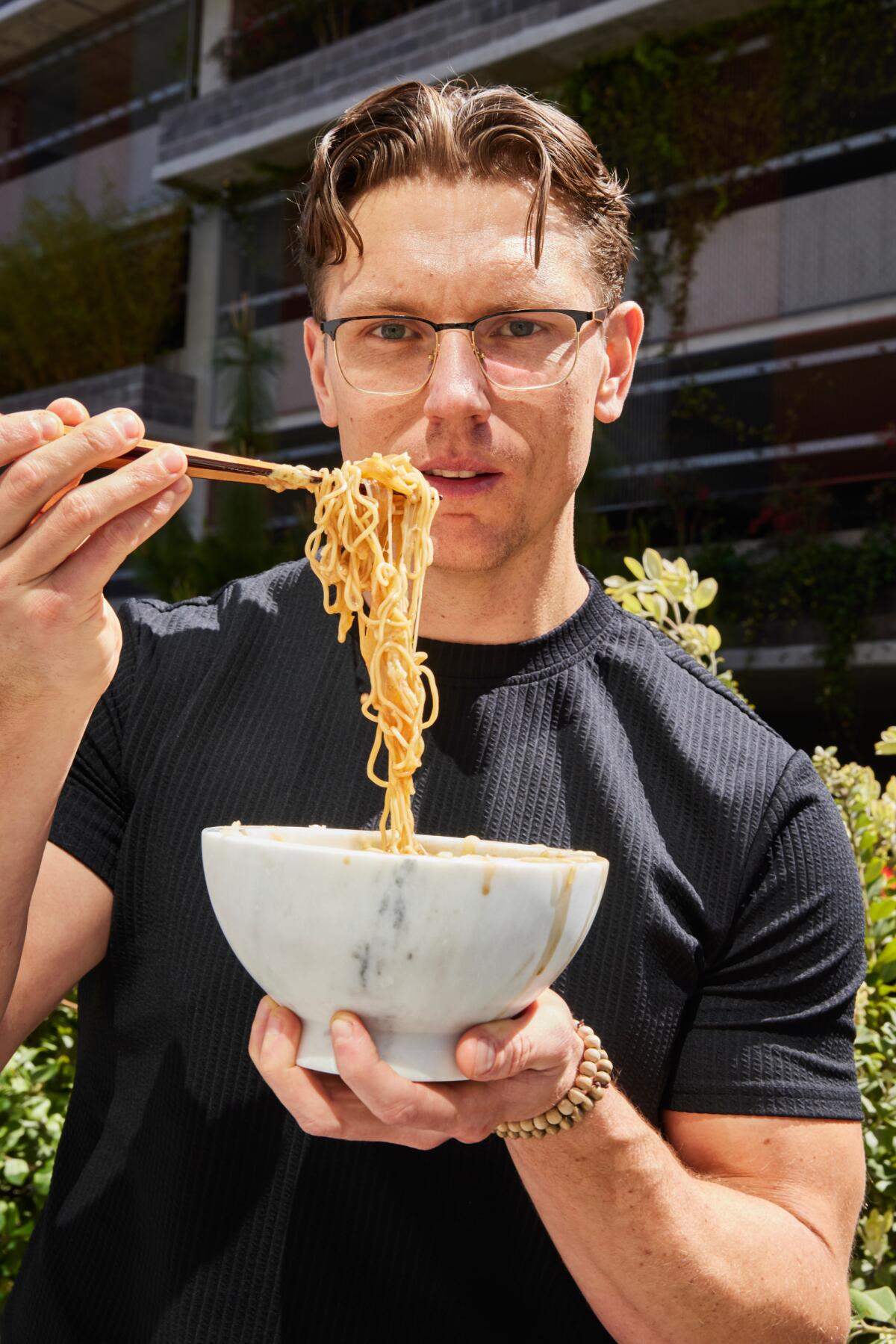 Chef Josh Scherer uses chopsticks to lift French onion ramen from a white bowl he holds in one hand.