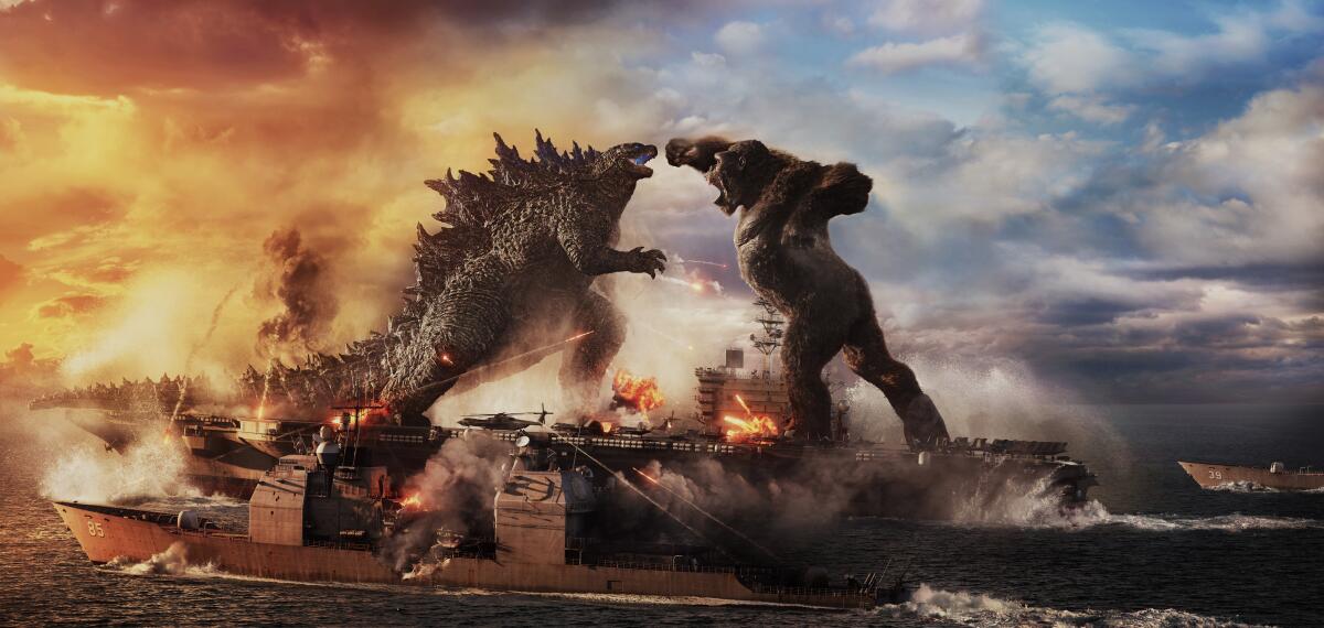 Two movie monsters face off in a harbor as fire and smoke rise.