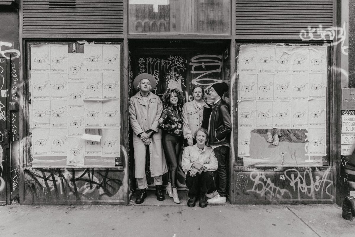 Five members of a rock band stand in front of a graffitied doorway