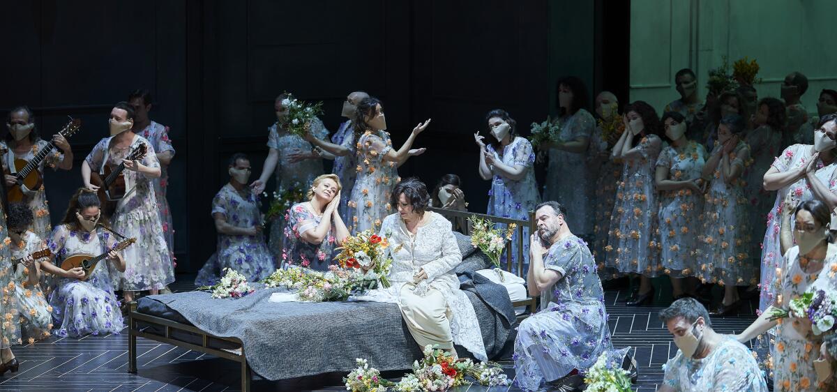 Performers in flowered gowns and strumming stringed instruments gather on a stage with a bed.