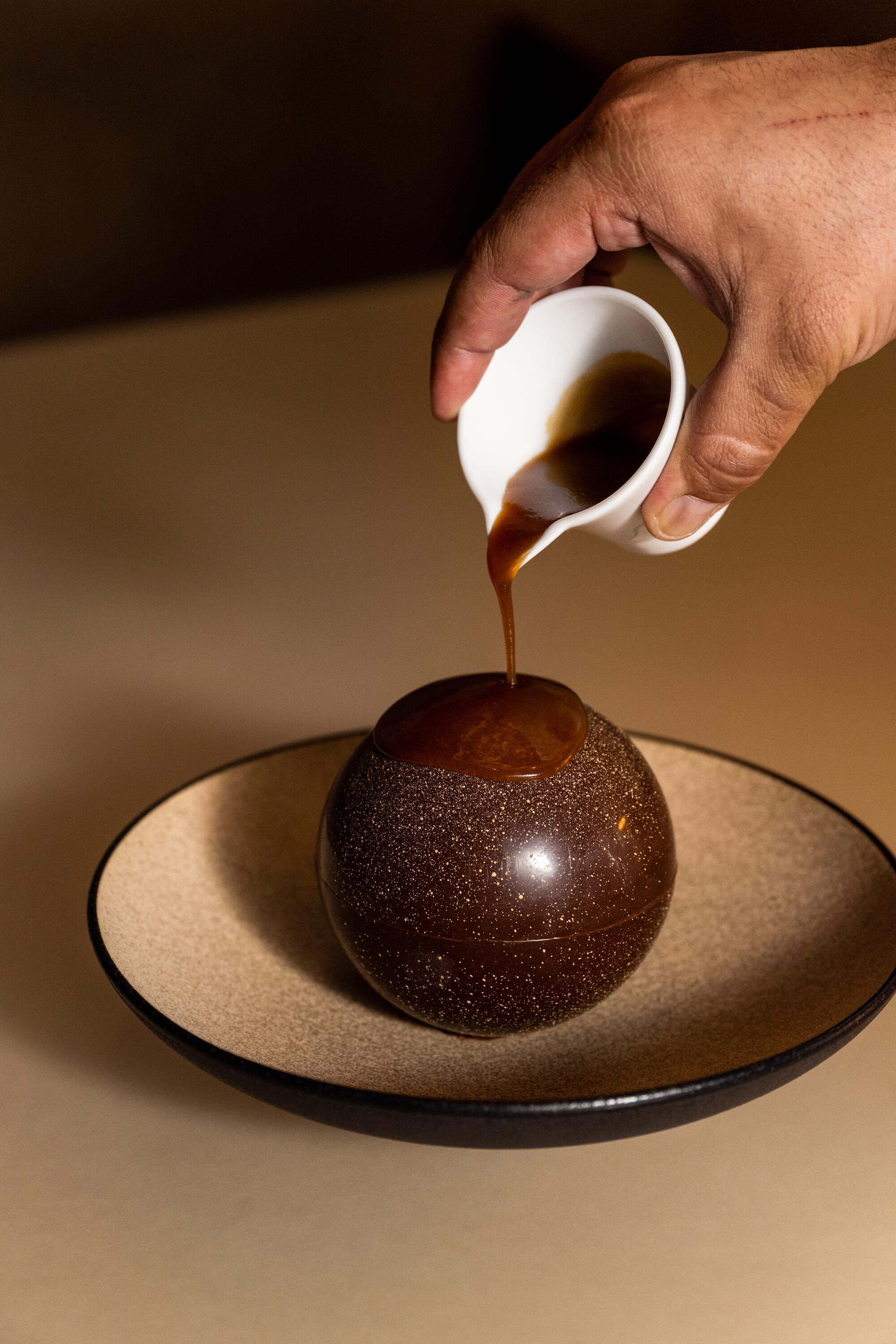 A hand pours caramel over a chocolate sphere