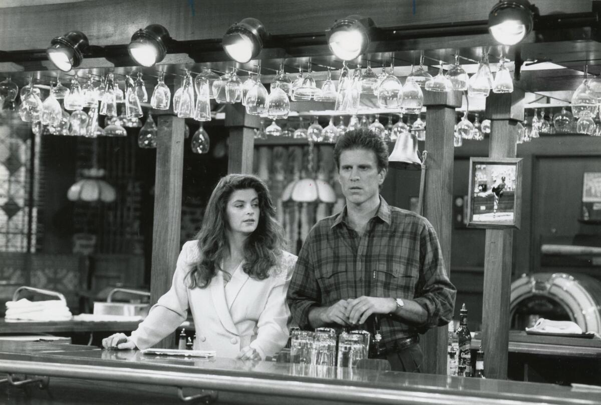 A man and a woman stand behind a bar on the TV series "Cheers."