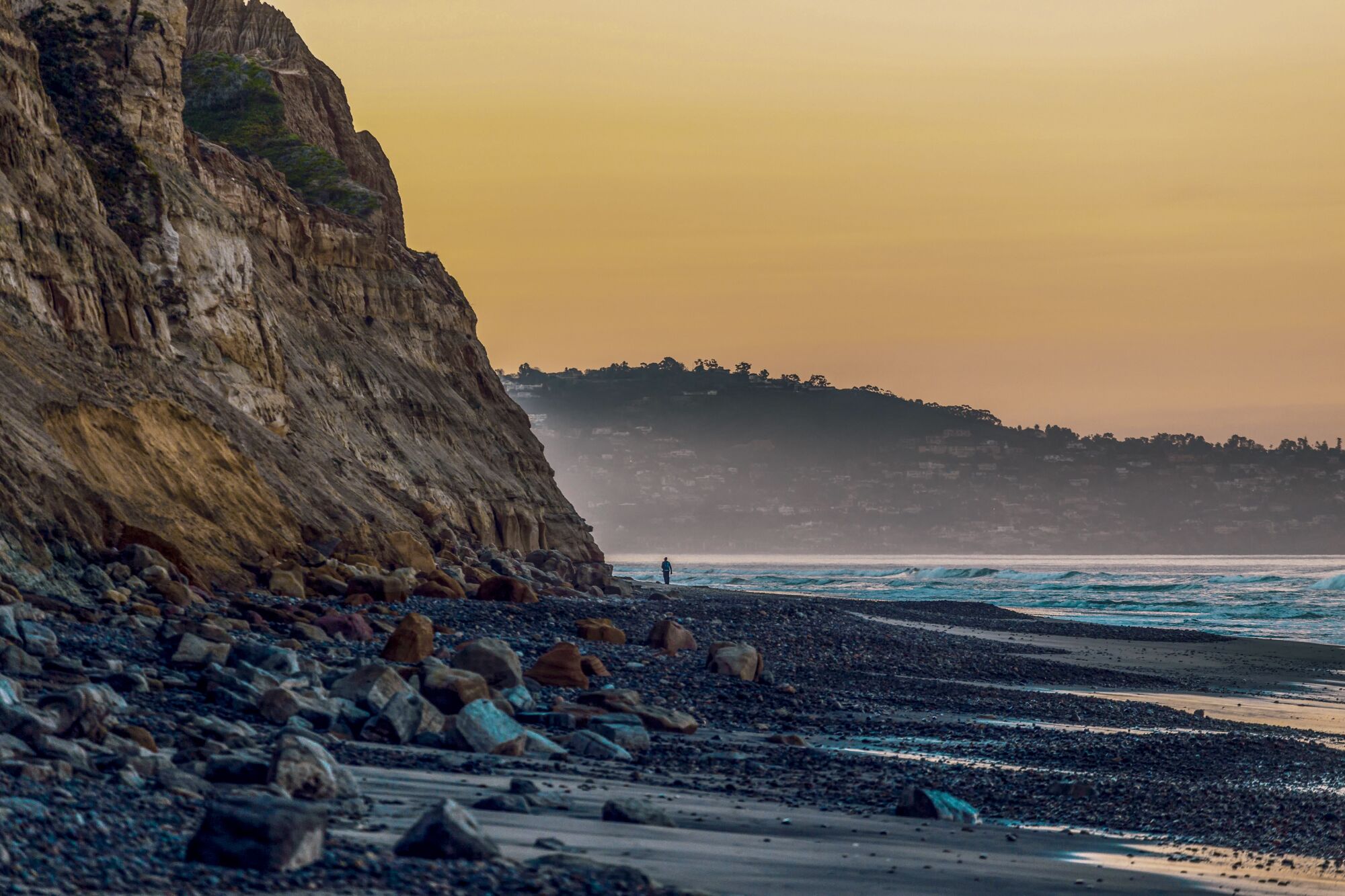 Distant silhouette of a person walking along the beach with rocky cliffs at Torrey Pines Beach.
