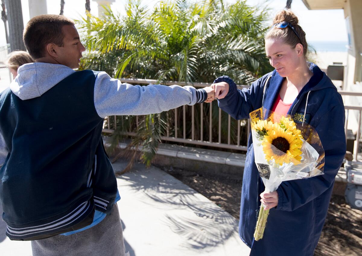 Two teems bump fists while standing outdoors. A girl at right is holding a bouquet of flowers. 