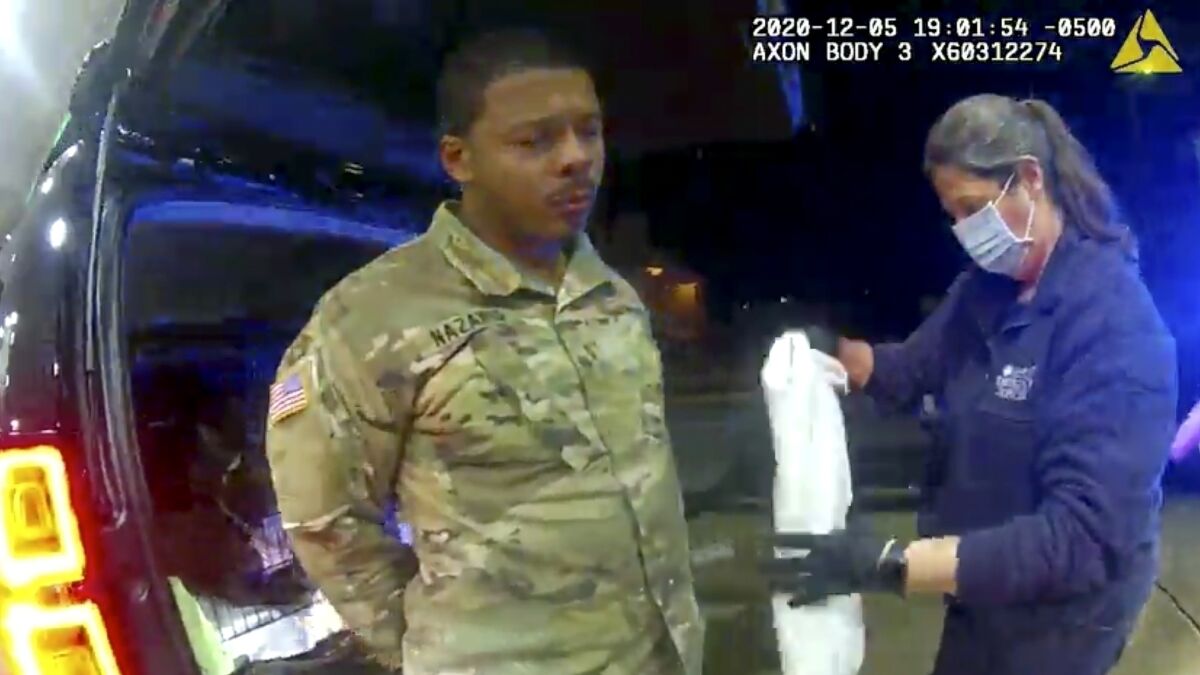 A police body camera image of a woman helping a man in a camouflage military uniform who stands behind a vehicle in the dark