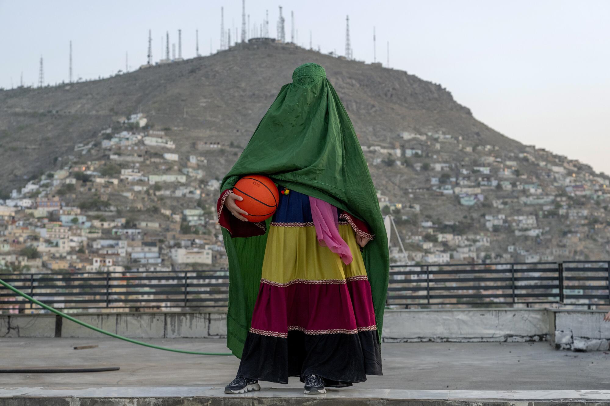 An Afghan woman wearing a green burqa and striped skirt poses with a basketball. 