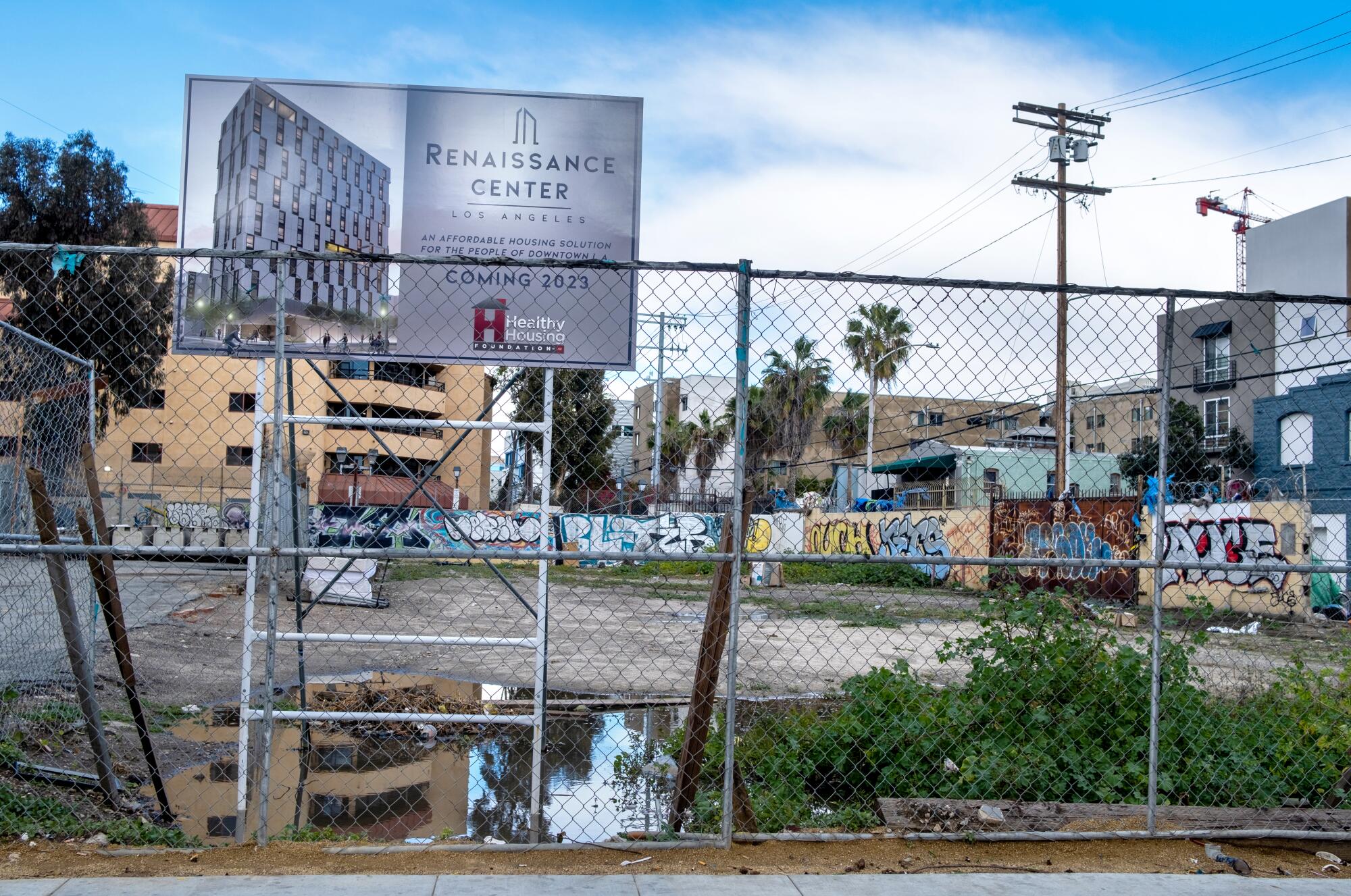 An empty lot behind a chainlink fence. Sign posted atop displays a grey modern building and title "Renaissance Center."