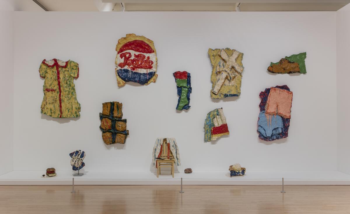 A series of sculptures on a wall, including one of a dress and another of a Pepsi sign.