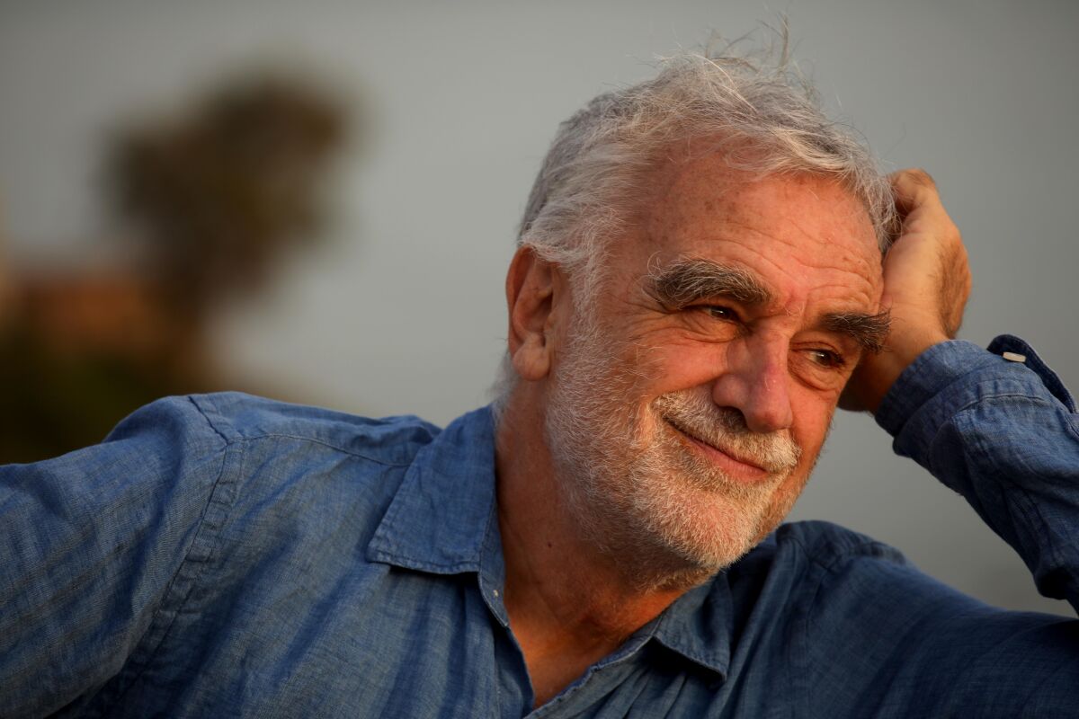 A head-and-shoulders portrait of a man with gray-white hair and short facial hair, leaning his head on his hand