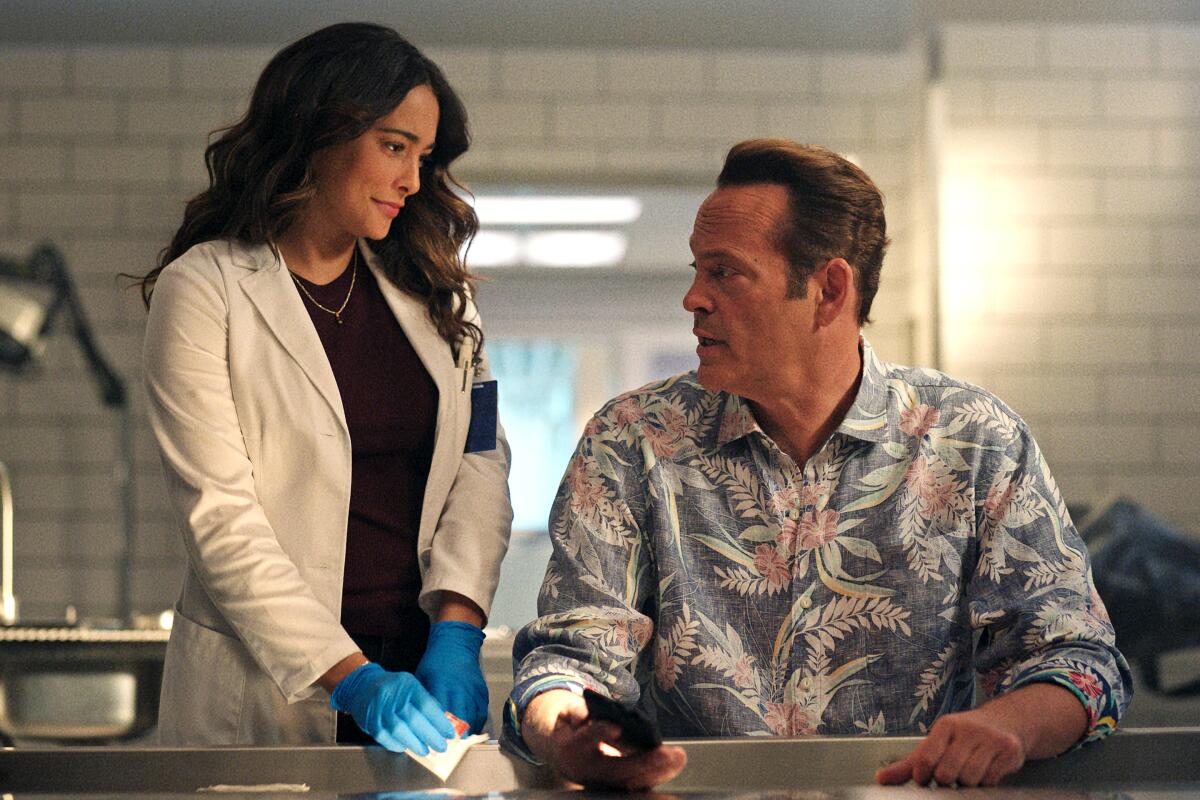 A woman with long brown hair and wearing a white lab coat stands next to a man in a Hawaiian shirt sitting a table. 