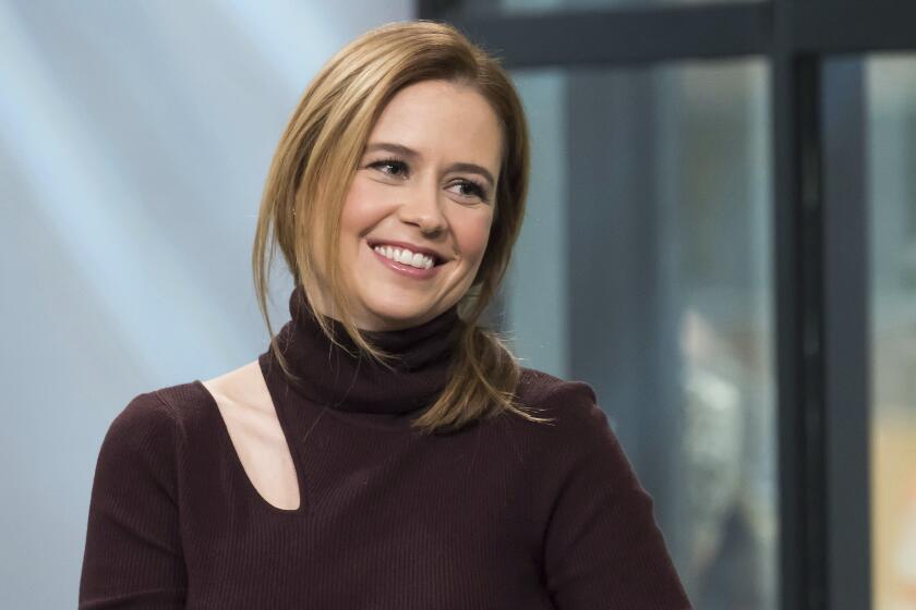 Jenna Fischer in a dark brown, mock-neck dress with a cut-out by her shoulder smiling and tilting her head to the right