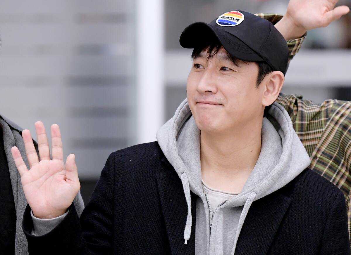 A man in a dark cap, gray hoodie and dark jacket smiles while waving
