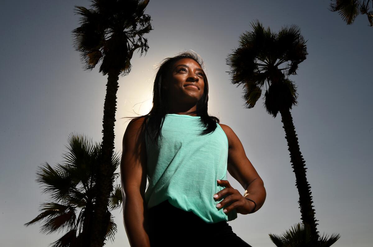 A portrait of Simone Biles, hand on hip and framed by palm trees.