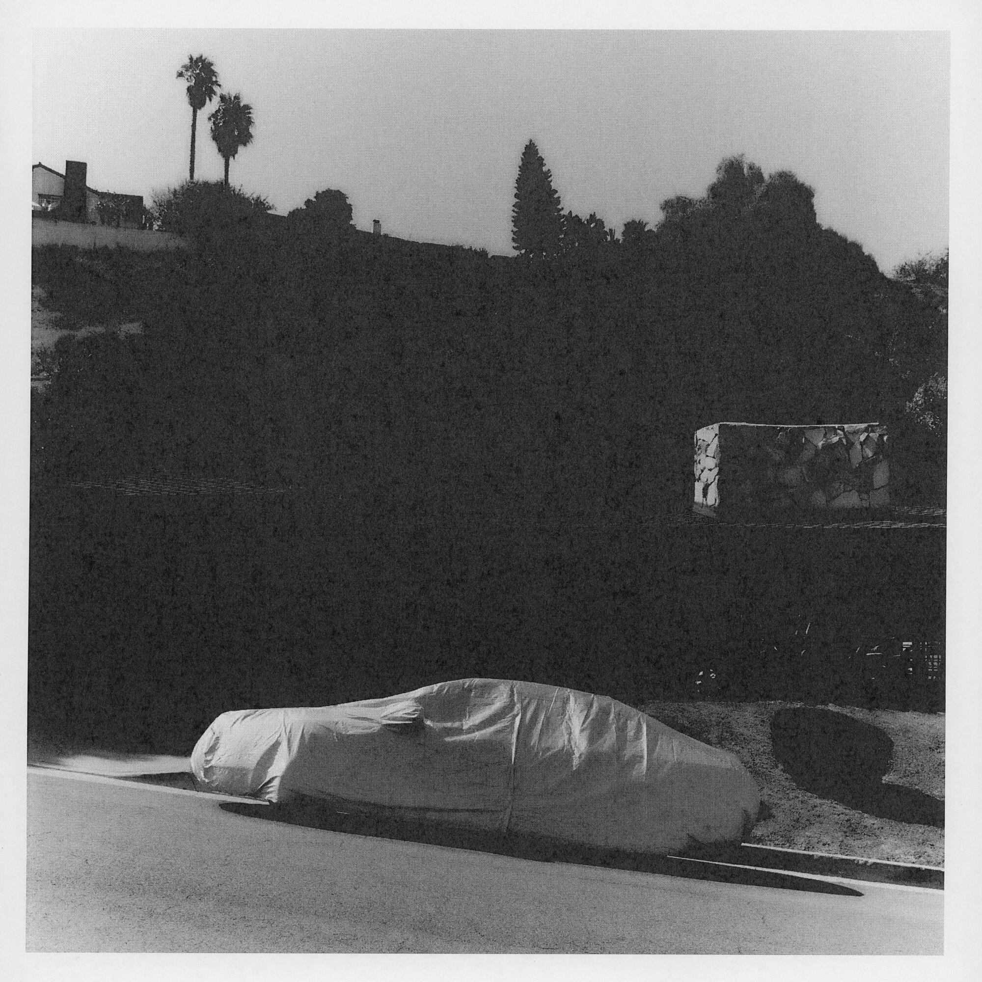 A black-and-white photo of a covered car parked on a sloping street.