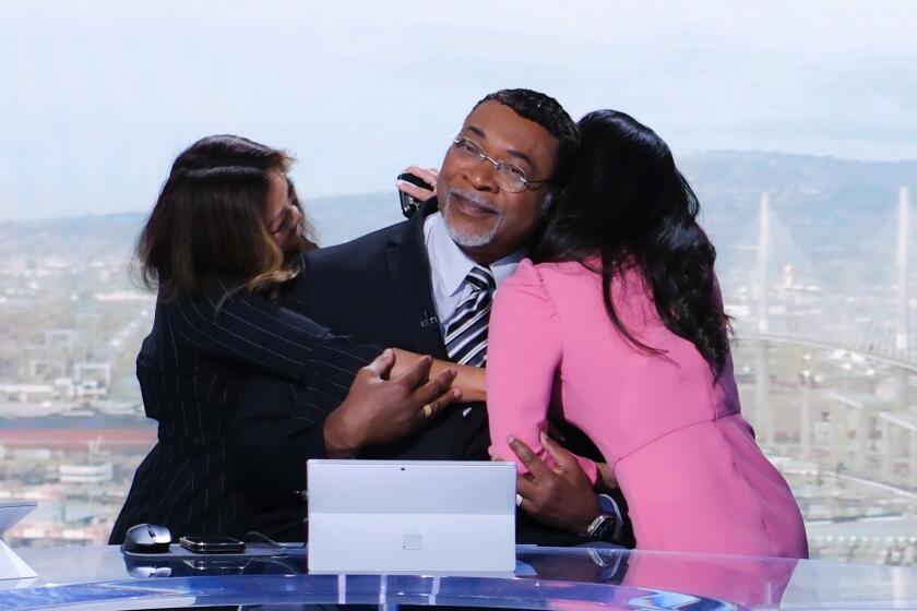 A man on a TV news set smiles as he gets hugs from two women colleagues
