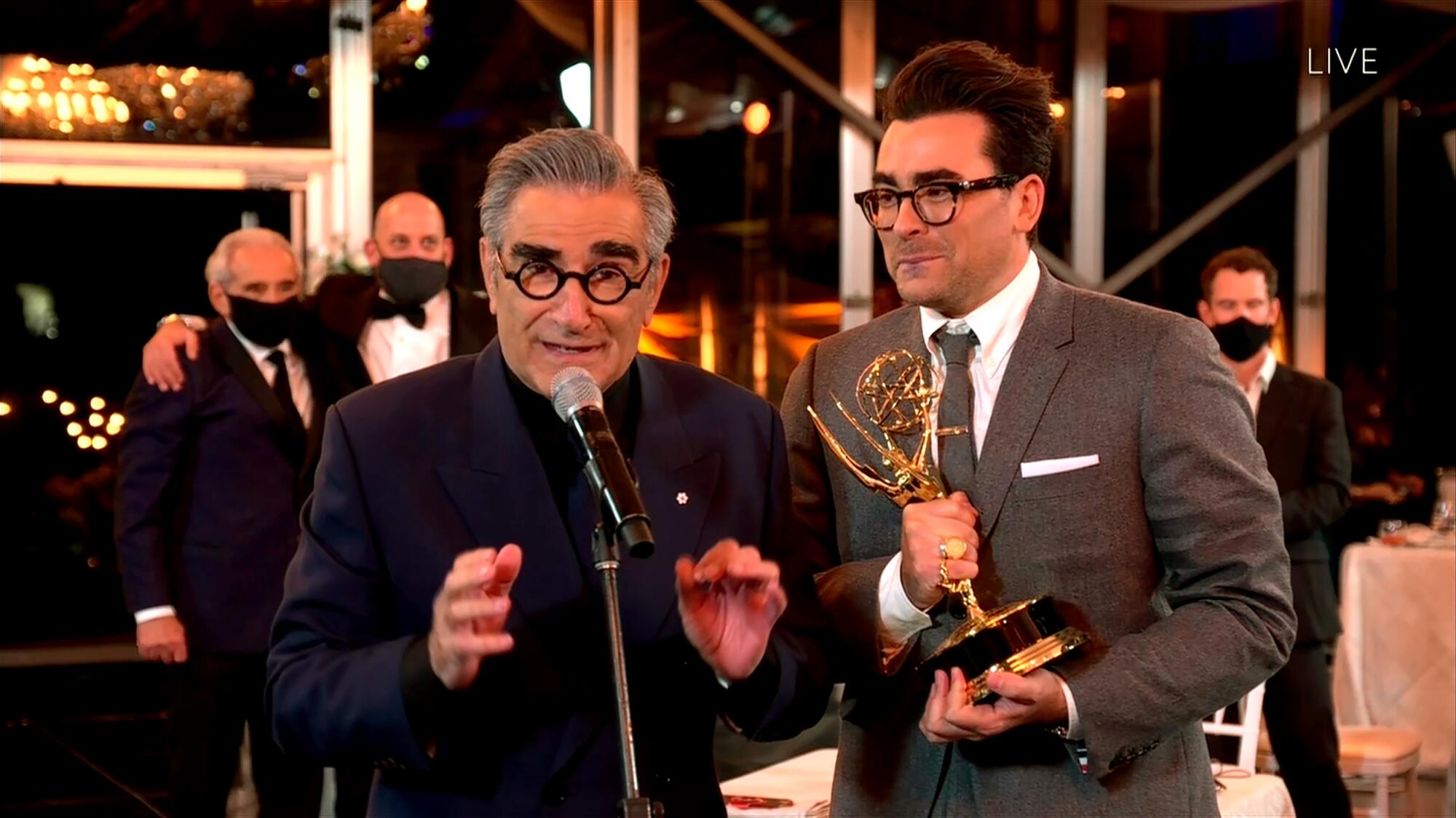 Eugene Levy in a black suit speaking into a microphone and Dan Levy standing next to him holding a golden statuette.