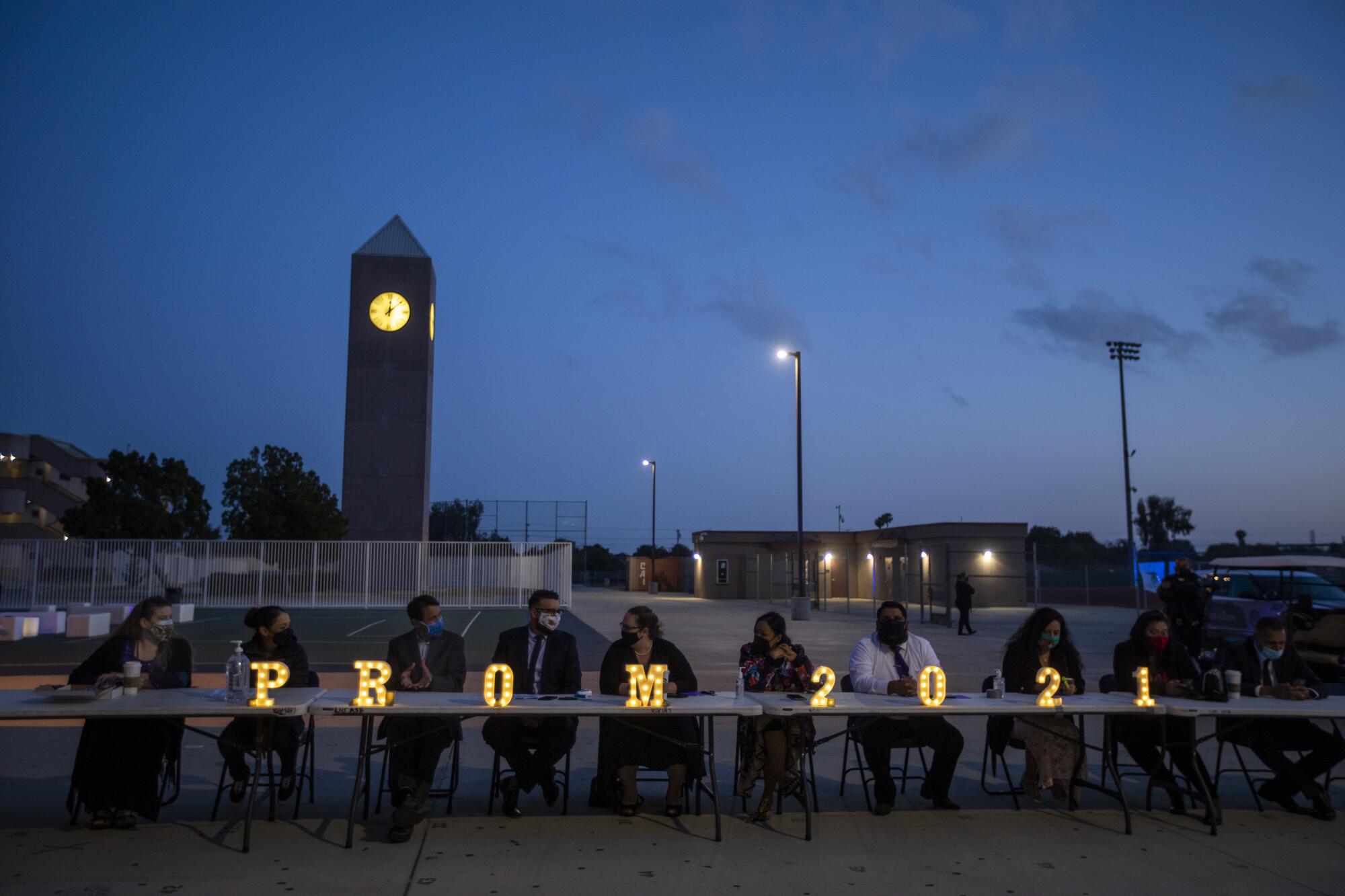 School faculty and staff sit at a table outside with light-up letters that read "Prom 2021"
