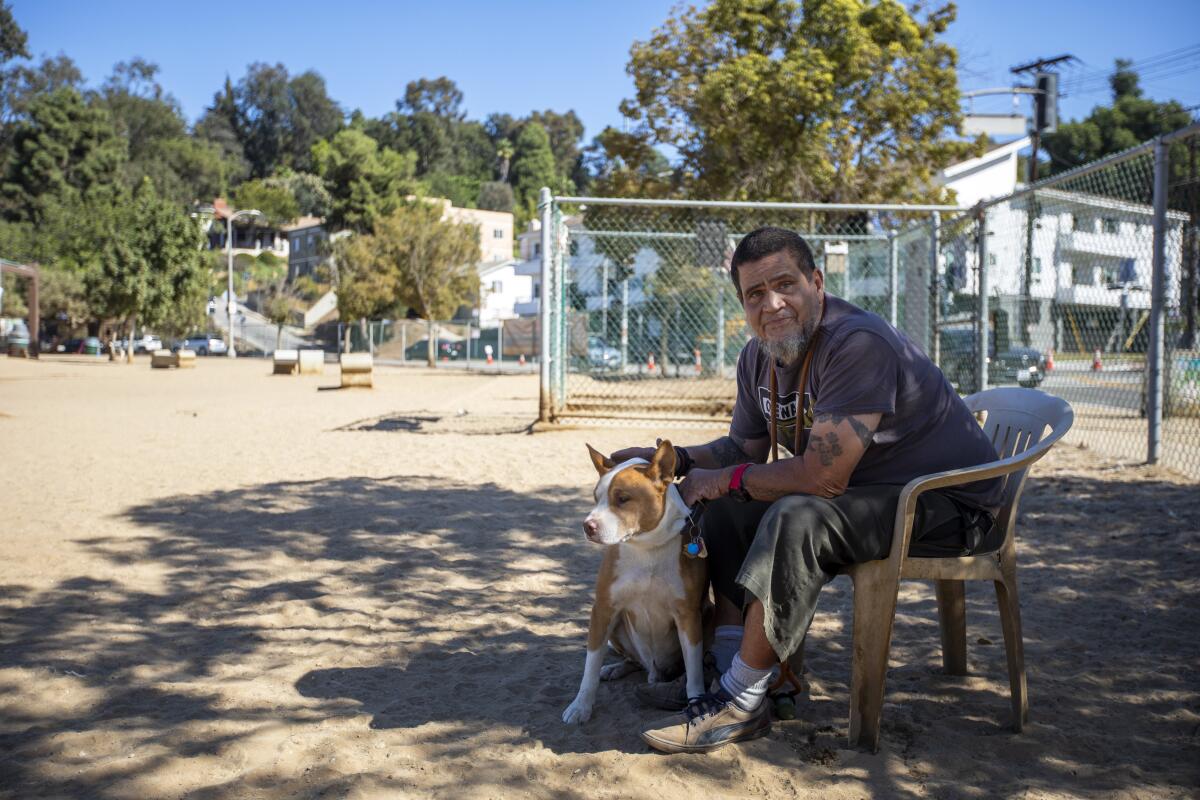 A man sitting on a chair in a dog park, holding on to a tan-and-white dog at his feet