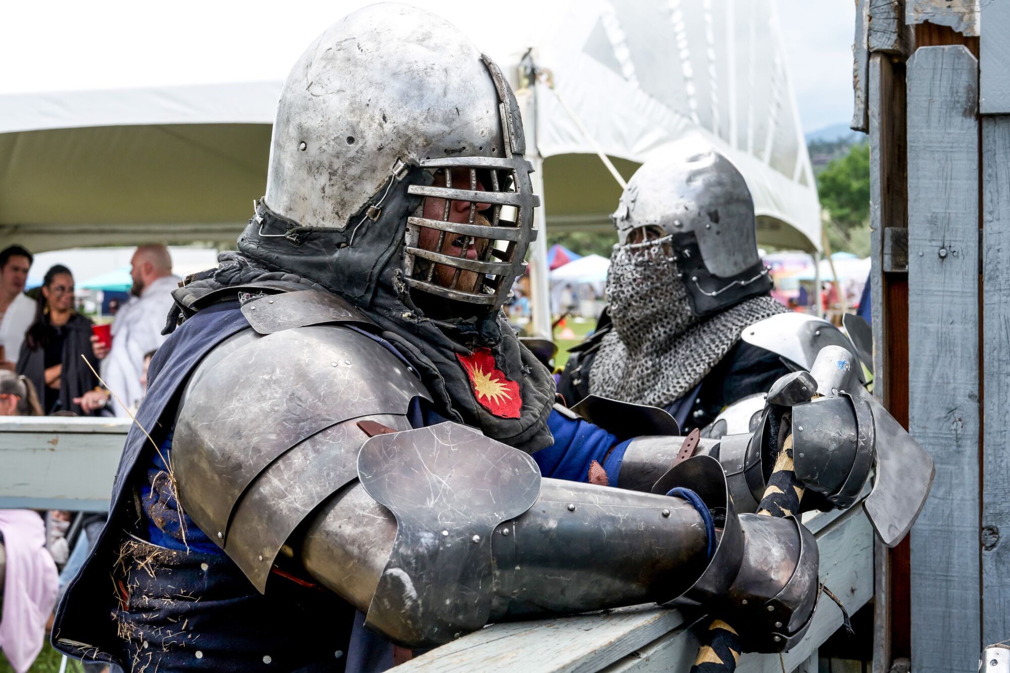 Participants in armor lean on a fence, waiting for a match to begin.