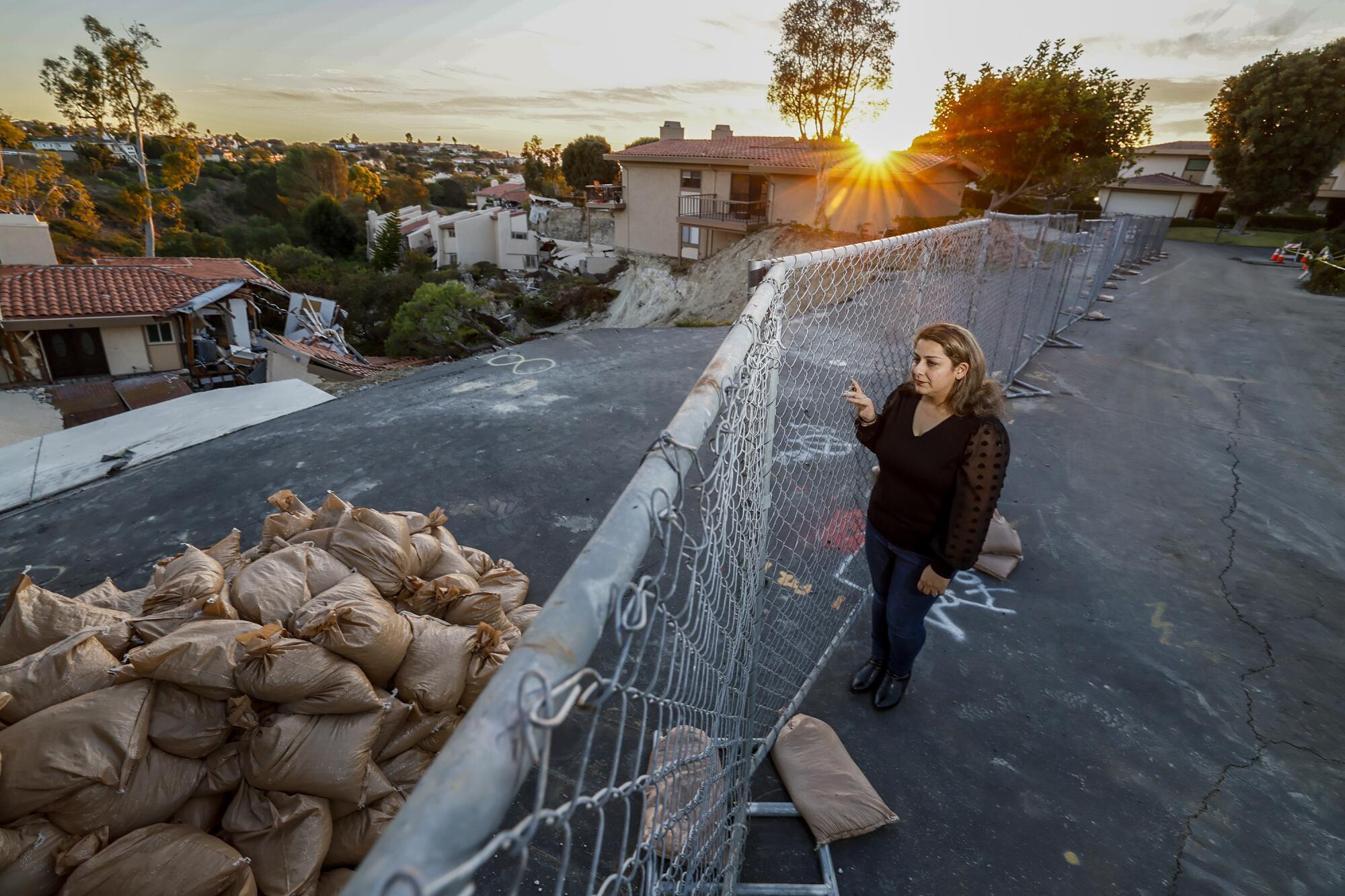 A woman stands next to a chain-link fence and sandbags, with damaged homes in the background.