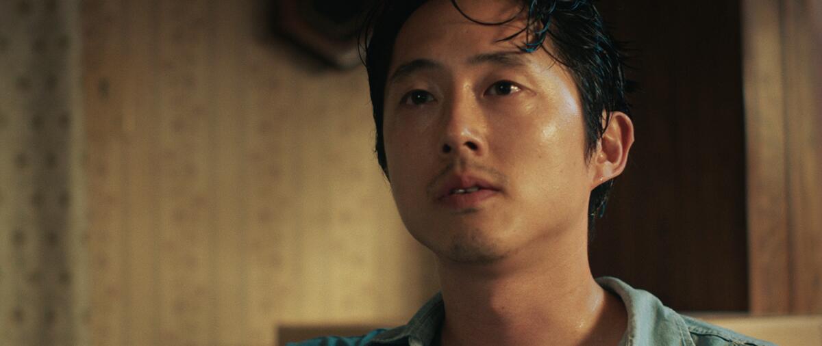Steven Yeun is looking up at someone, his face blank. There is a patterned wallpaper behind him.