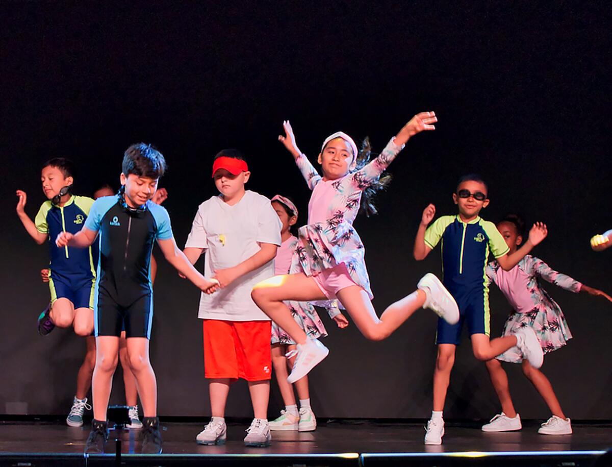 Kids in wetsuits perform onstage. A girl in pink jumps and poses in the center.