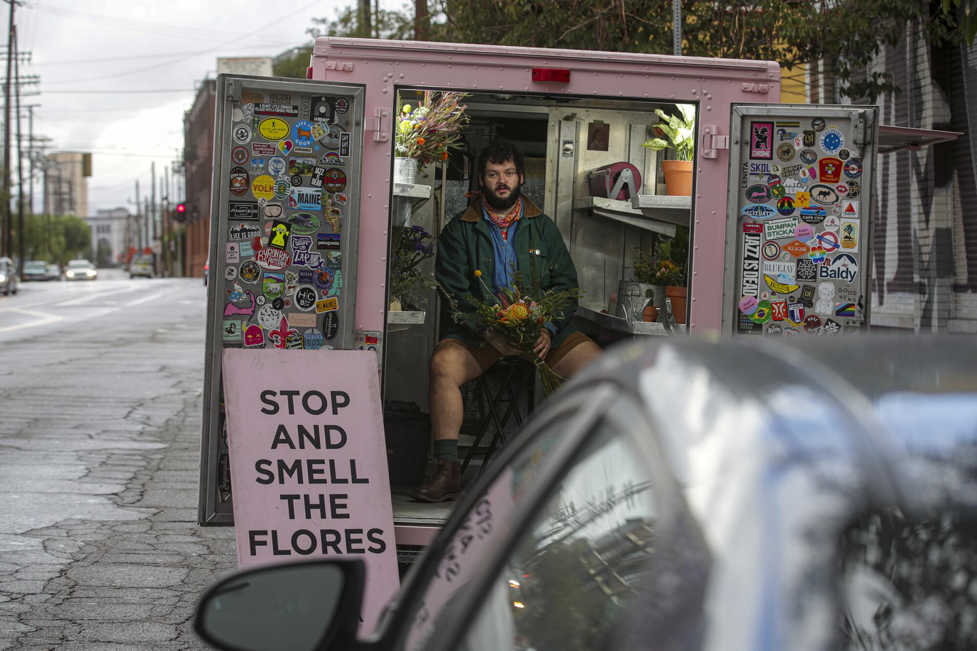 A man sits in the back of an open, pink truck next to a sign reading "Stop and Smell the Flores."