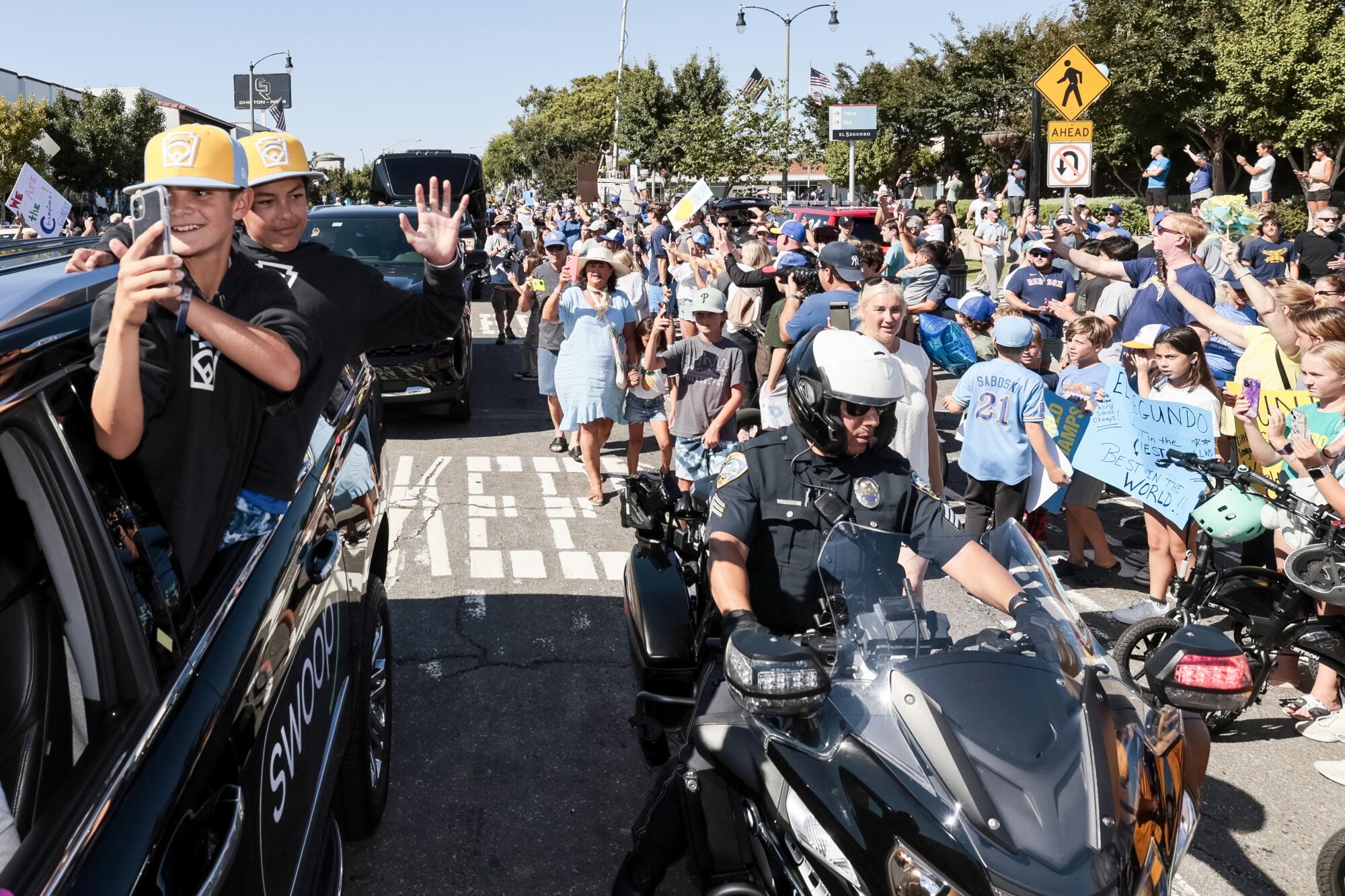 A cheering crowd surrounds black SUVs holding boys in baseball caps. A motorcycle officer polices the crowd.