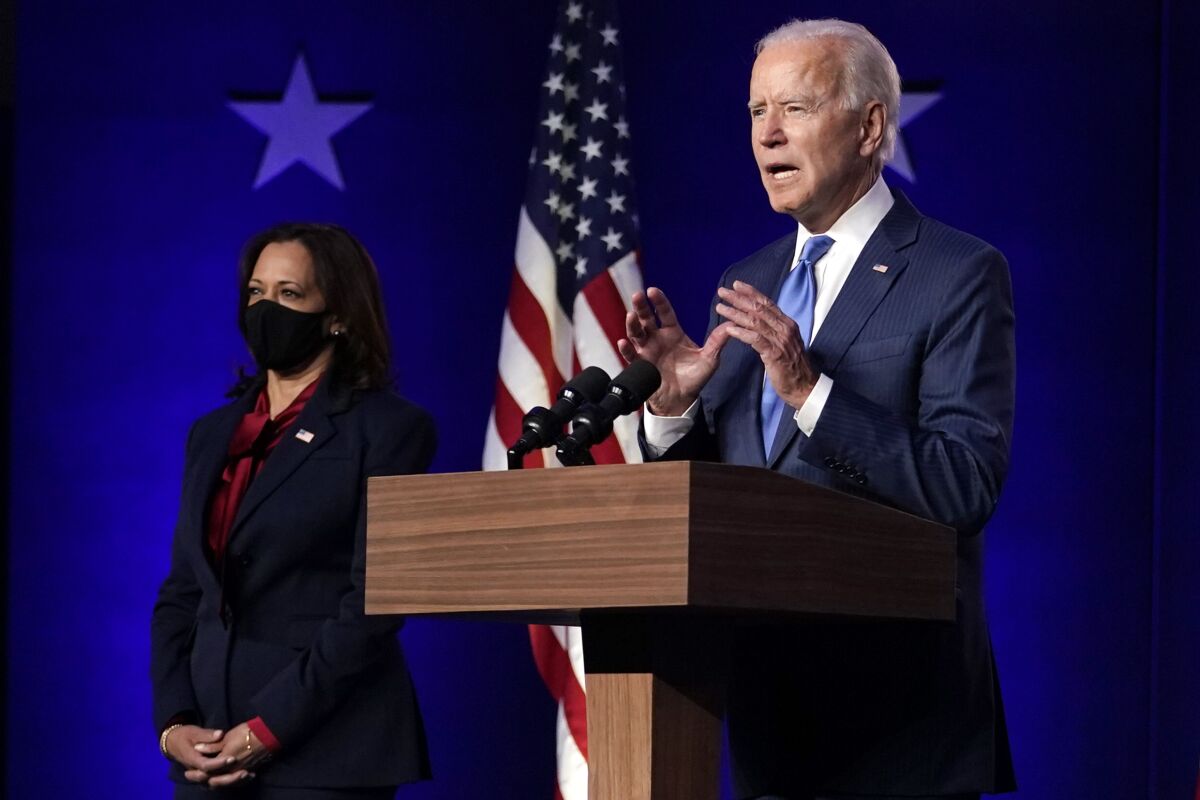 Joe Biden gestures with both hands as he speaks at a lectern with Kamala Harris, wearing a mask, standing nearby.