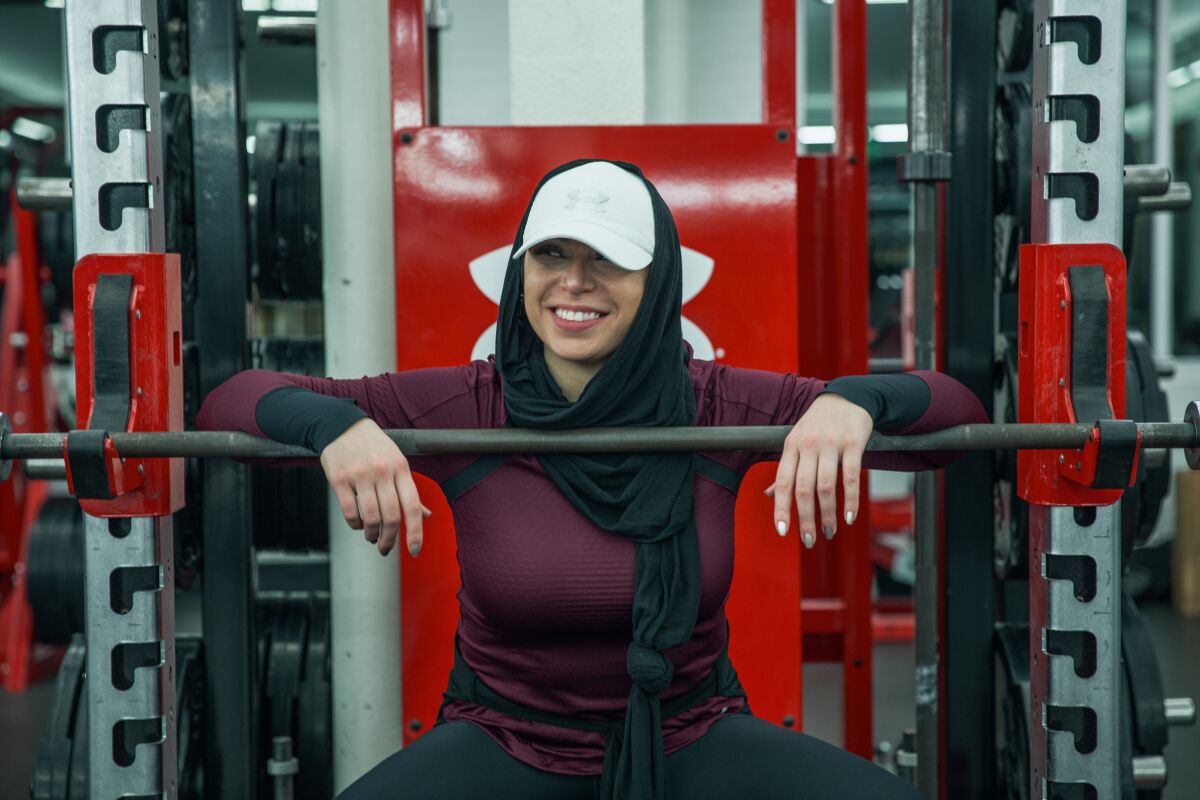 A woman in a hijab and workout clothes poses with weightlifting equipment