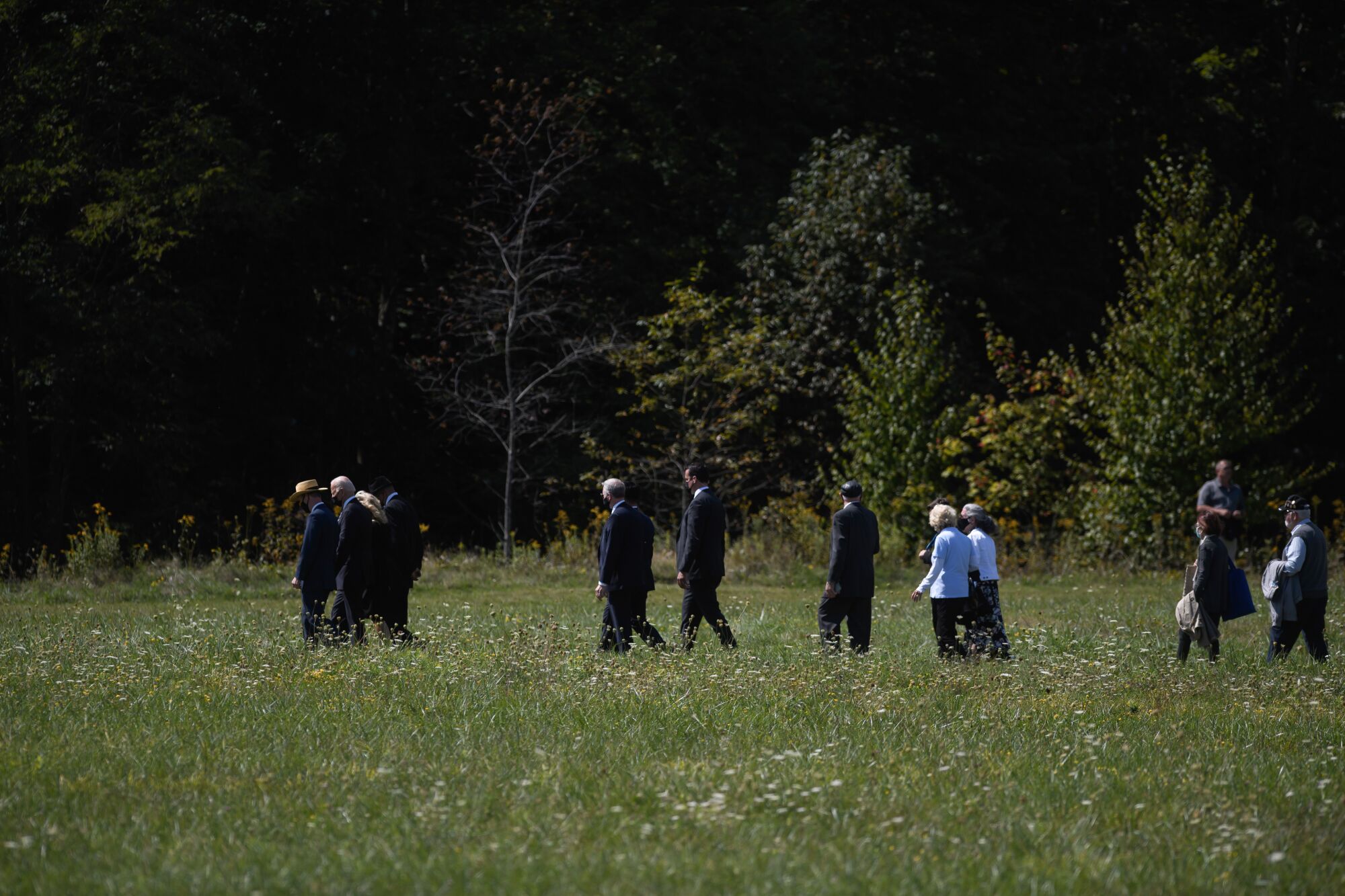 Several people in suits and dresses walk across a grassy field.
